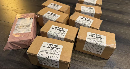 Multiple reused cardboard shipping boxes with ‘I am a box with a history’ labels, part of TimPrintDreams’ Box Revival eco-friendly packaging project, placed on a wooden floor alongside one pink parcel.